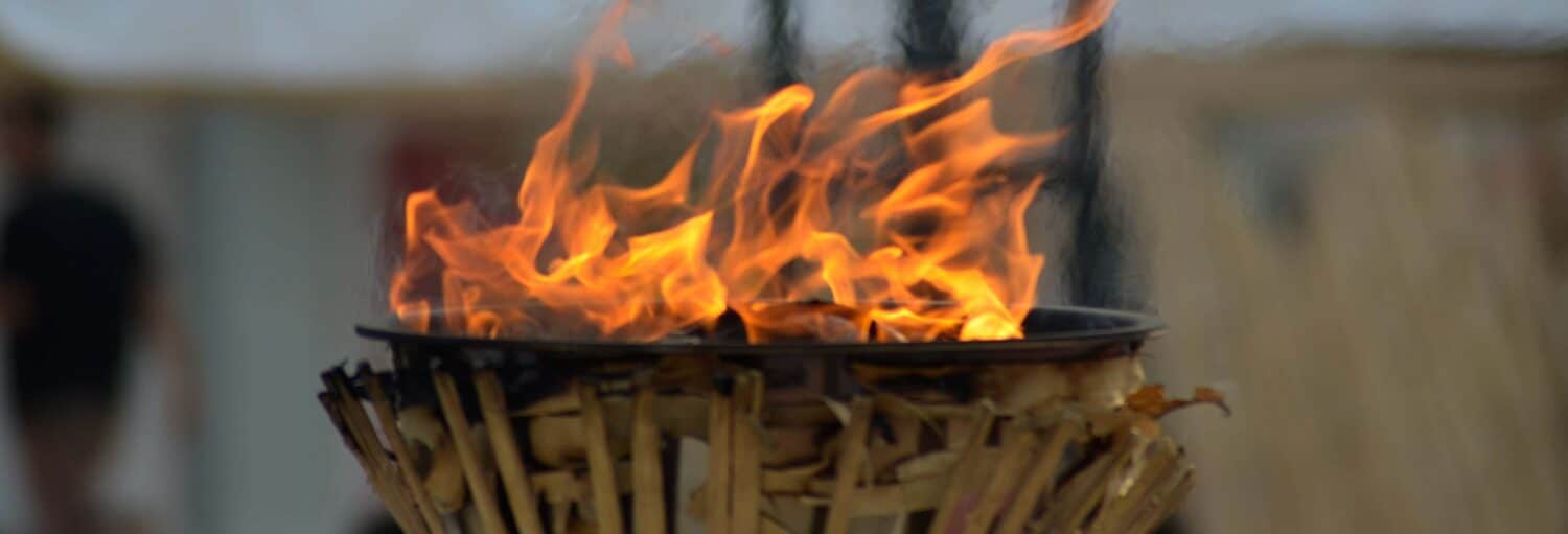 Um close-up de uma tocha externa feita de madeira e metal com uma chama laranja brilhante acesa. A imagem ilustra o post sobre Ingresso do Museu Olímpico de Atenas.