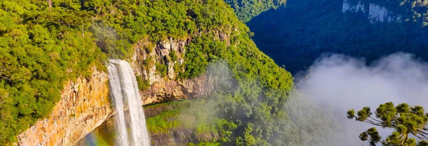 Uma cachoeira alta cai em cascata sobre um penhasco rochoso cercado por uma densa floresta verde, com a névoa e a luz do sol criando um leve arco-íris. - Ingresso do Parque do Caracol