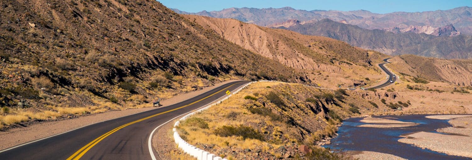 Uma estrada sinuosa e pavimentada passa ao lado de um rio raso por colinas secas e rochosas e montanhas distantes sob um céu claro no Excursão de alta montanha pelos Andes em grupo reduzido