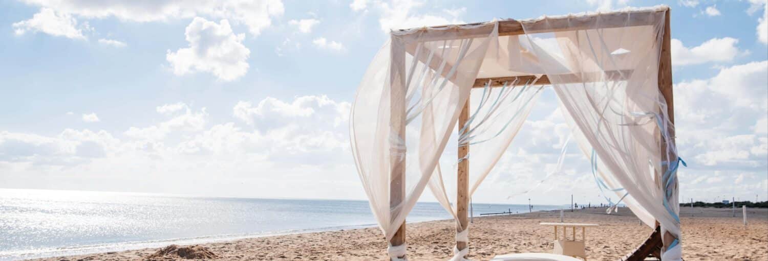 Um dossel de madeira com cortinas brancas fica em uma praia de areia perto do mar calmo sob um céu parcialmente nublado. Imagem para ilustrar post sobre a excursão ao Bora Bora Beach.