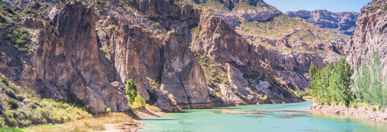 Um rio azul-turquesa flui por um cânion rochoso com penhascos íngremes e vegetação verde esparsa sob um céu claro durante a Excursão ao Cânion do Atuel e San Rafael