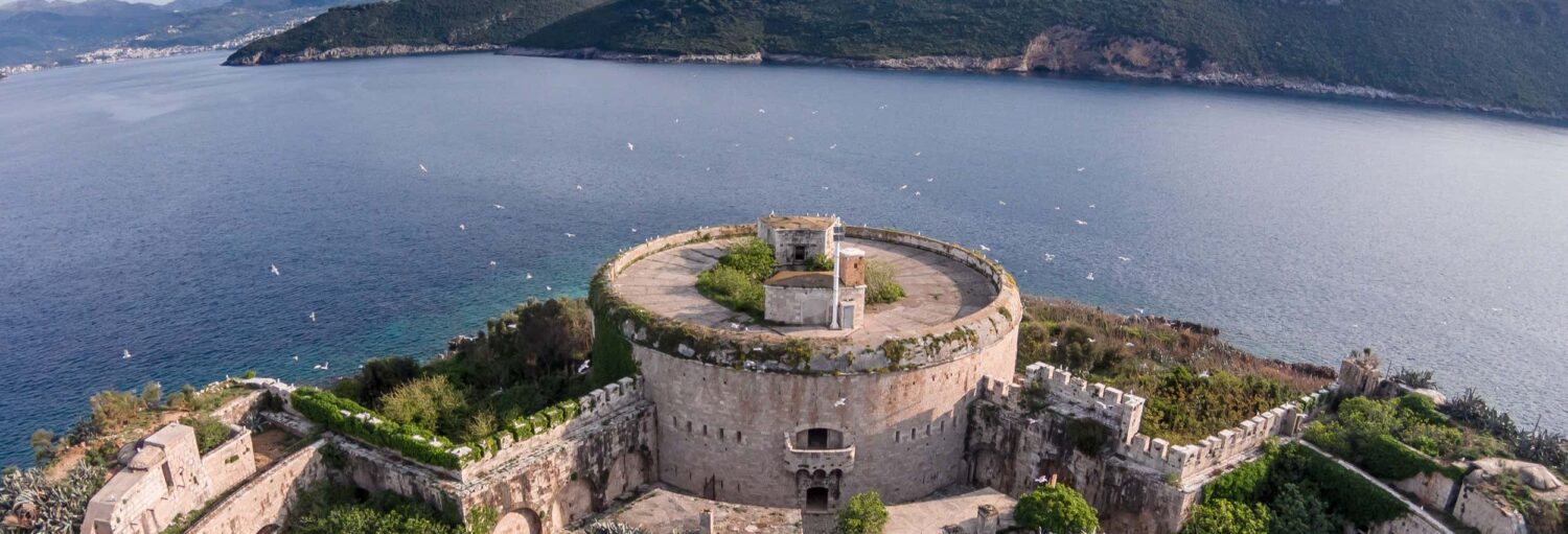 Uma fortaleza redonda de pedra com vista para a água azul e colinas distantes, cercada por vegetação verde e antigas paredes de pedra, para representar Excursão à Gruta Azul e Nossa Senhora das Rochas de barco.