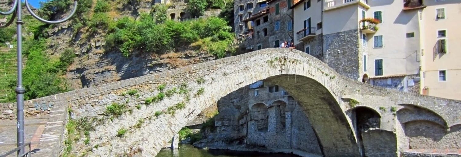 Uma ponte de pedra em arco atravessa um rio ao lado de prédios antigos construídos em uma encosta coberta de vegetação. - Excursão a San Remo e Dolceacqua