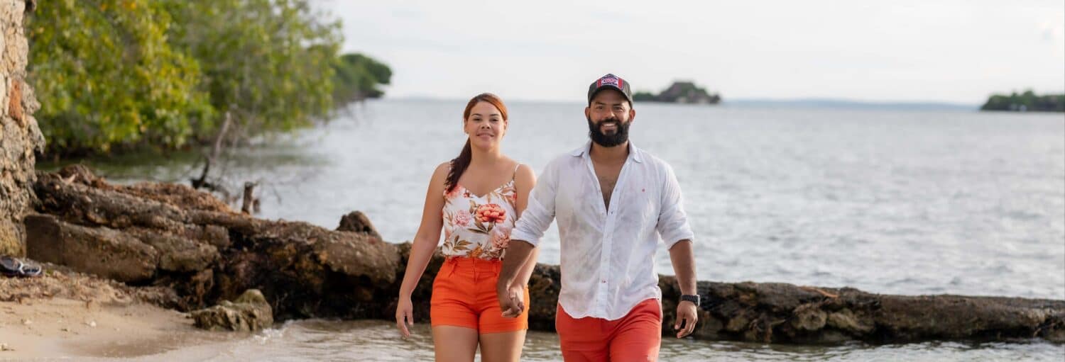 Um homem e uma mulher de mãos dadas caminham em uma praia de areia com água e árvores ao fundo. Imagem para ilustrar post sobre a excursão à Ilha de Cocoliso.