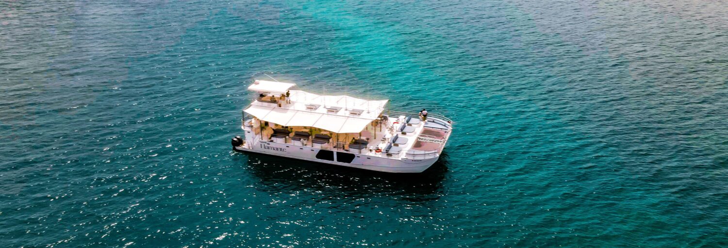 Um barco branco com cobertura flutua em uma água azul clara, visto de cima em um dia ensolarado. Imagem para ilustrar post sobre a excursão à Isla Grande.