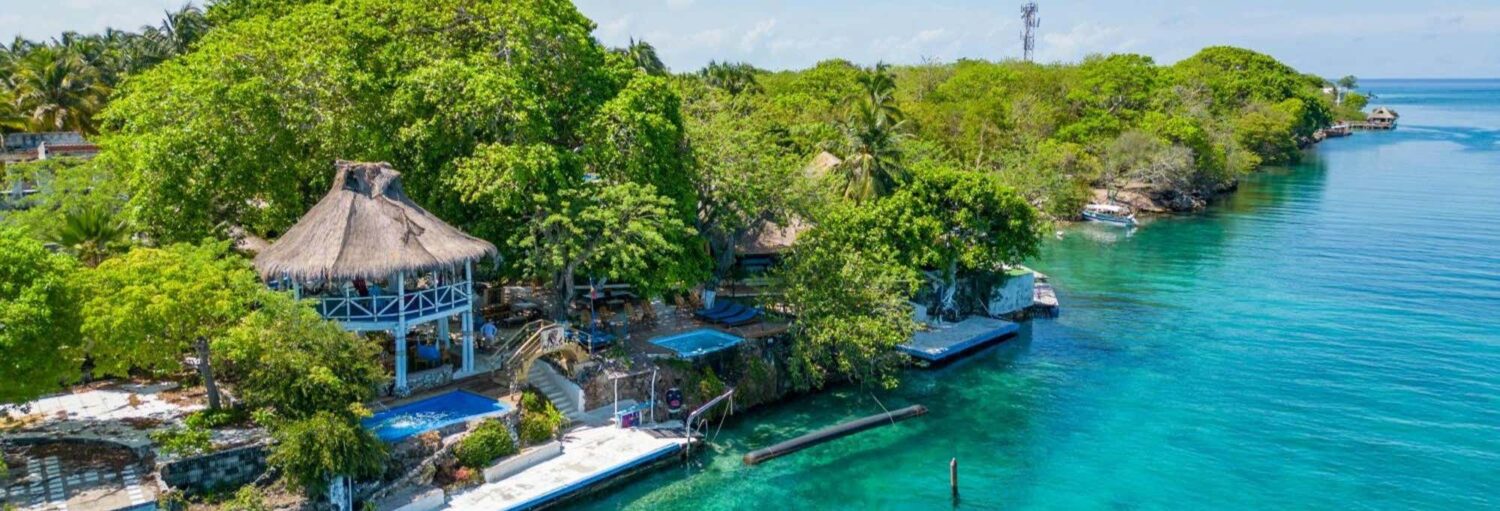 Um prédio com telhado de palha e piscinas ficam entre as árvores ao lado de água azul clara em uma costa tropical exuberante. Imagem para ilustrar post sobre a excursão ao Isla Lizamar Beach Club.