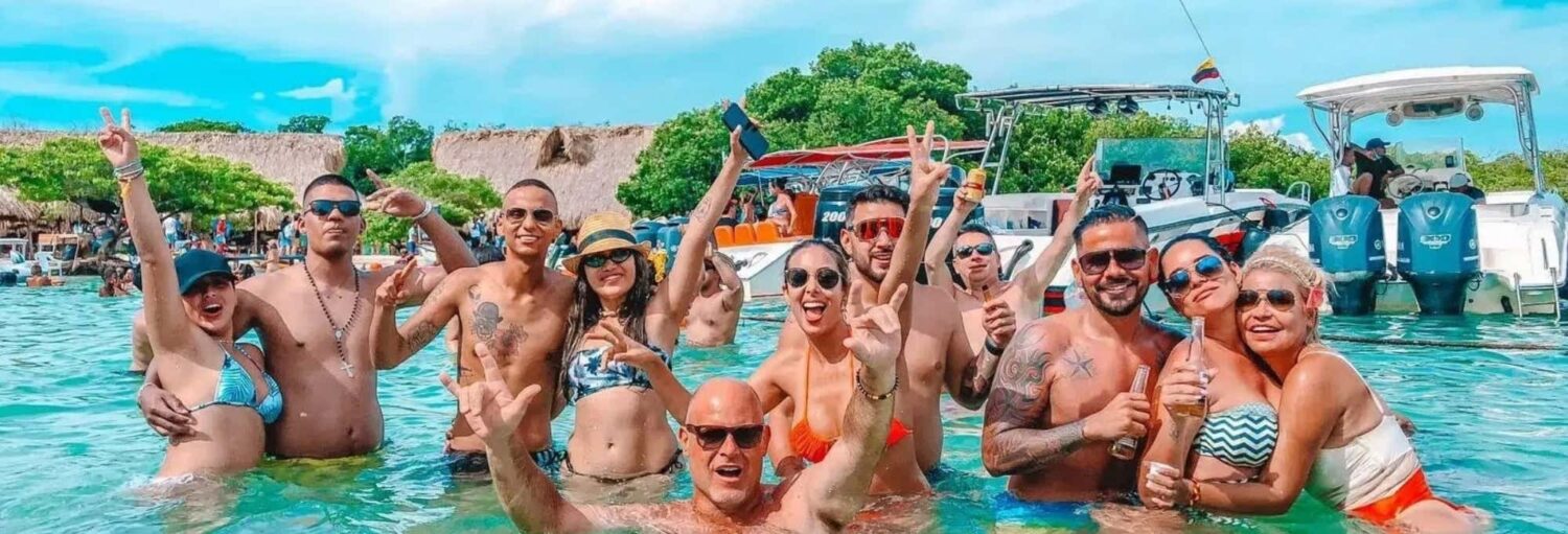 Um grupo de pessoas em trajes de banho sorrindo e posando juntas em águas rasas com barcos e árvores ao fundo. Imagem para ilustrar post sobre a excursão às ilhas do Rosário para adultos.