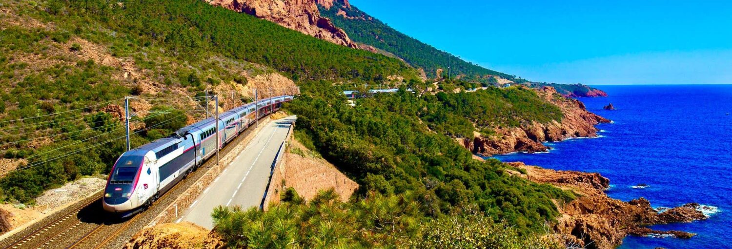 Um trem de alta velocidade viaja ao longo de uma ferrovia costeira com colinas e vegetação de um lado e o mar azul do outro. - Excursão ao Maciço de l'Estérel de trem