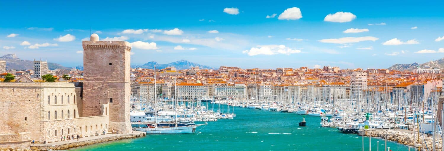 Vista de uma marina com muitos barcos, uma torre de pedra histórica e uma paisagem urbana ao fundo, sob um céu azul com nuvens. - Tour de Marselha para cruzeiros
