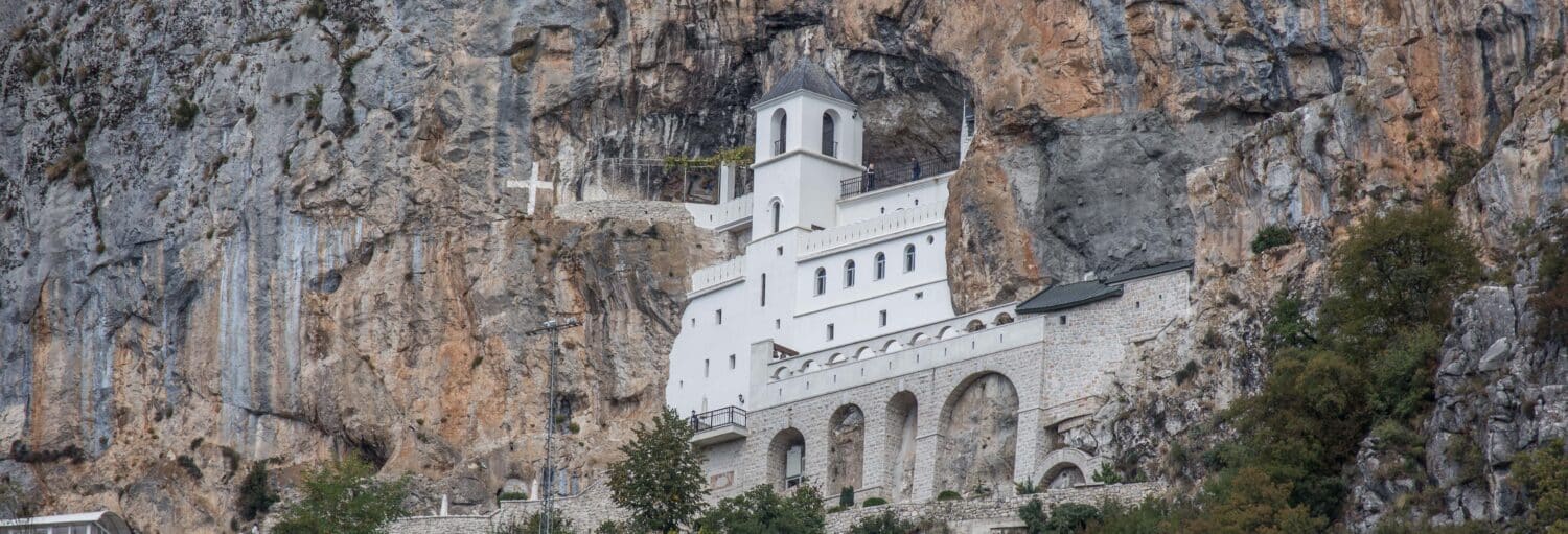 Mosteiro branco construído em um penhasco íngreme e rochoso, com uma grande cruz e algumas árvores nas proximidades, na Excursão ao mosteiro de Ostrog.