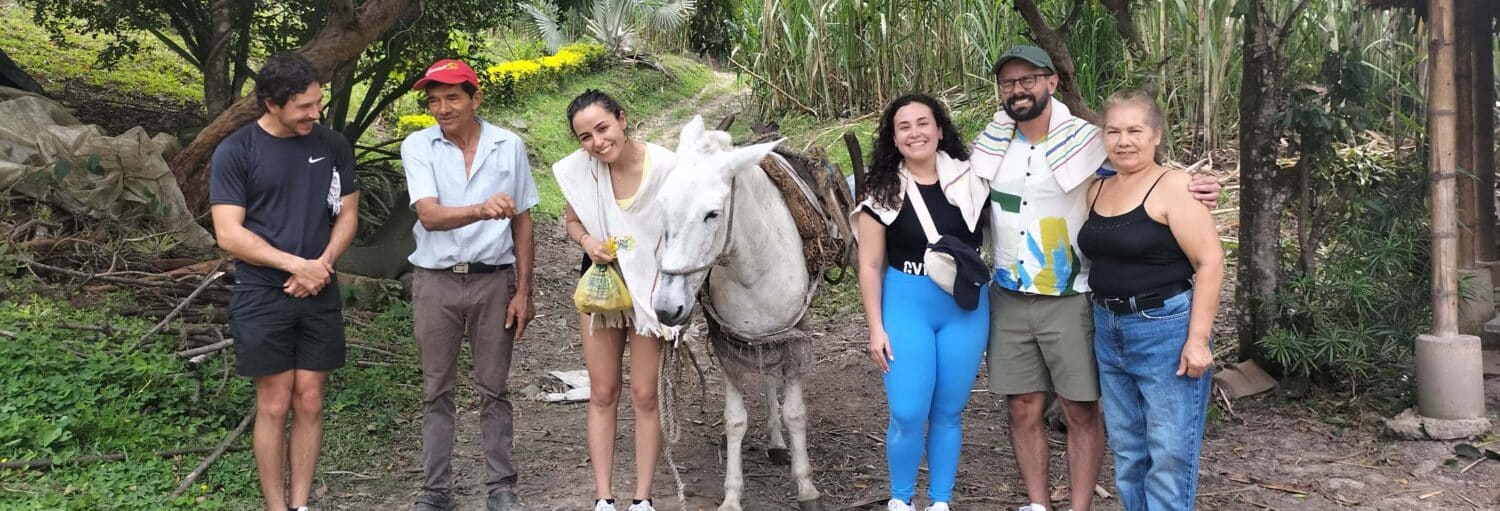 Seis pessoas estão ao ar livre com um burro branco, cercadas por árvores e vegetação, todas de frente para a câmera. A imagem ilustra o post sobre Tour por Nocaima, La Vega e fazenda El Bambú.