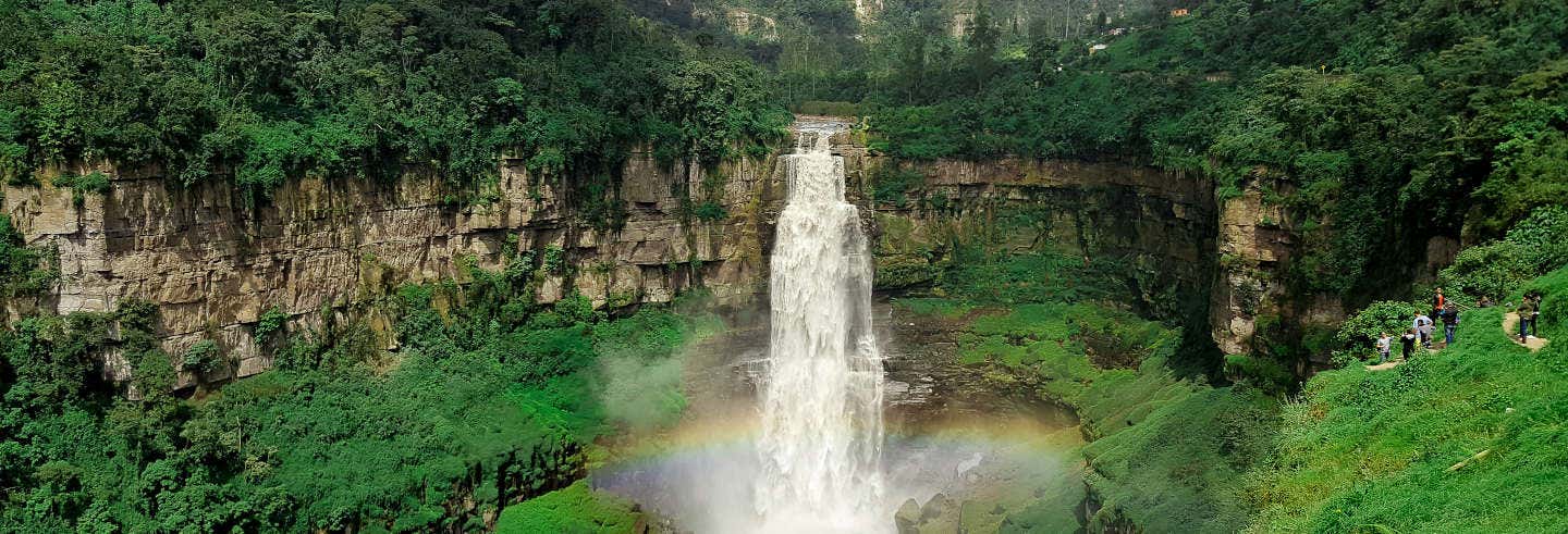 Uma alta cachoeira que deságua em um vale verde em uma excursão à catarata de Tequendama, com um arco-íris brilhando na base da floresta.