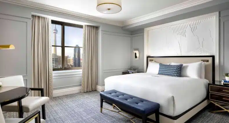 Quarto do hotel Fairmont Royal York moderno com uma cama grande, banco azul, mesa com cadeiras, janela com vista para a cidade e decoração neutra.