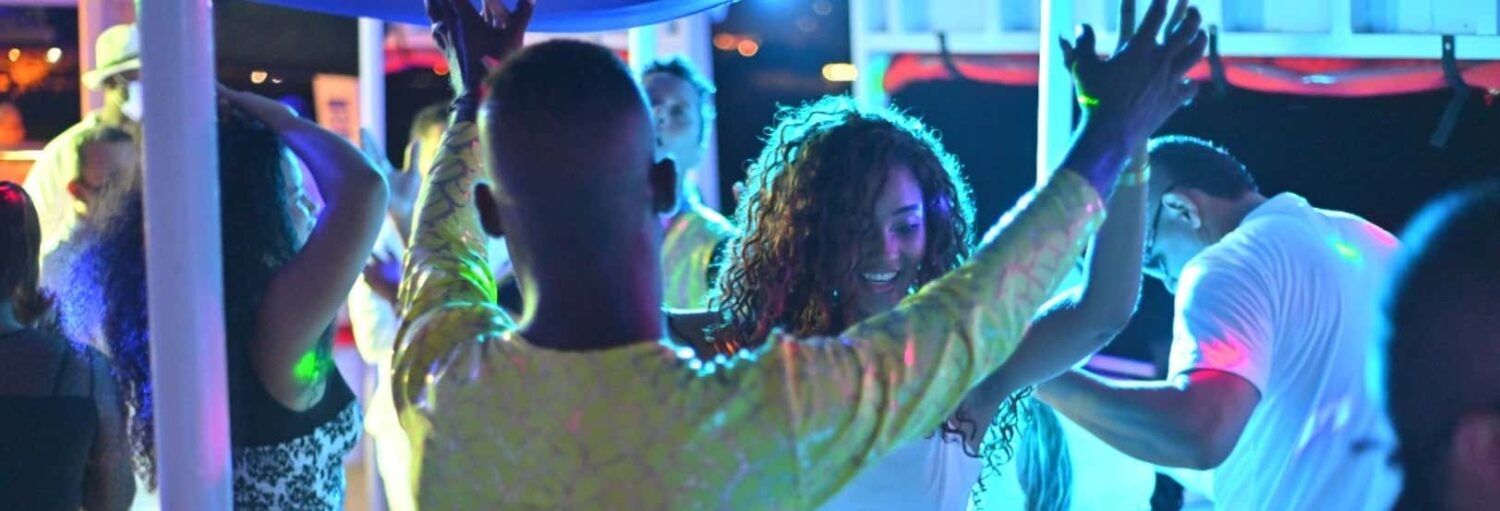 As pessoas estão dançando sob luzes coloridas em uma festa, com algumas levantando os braços e sorrindo. Imagem para ilustrar post sobre a festa caribenha de barco por Cartagena.
