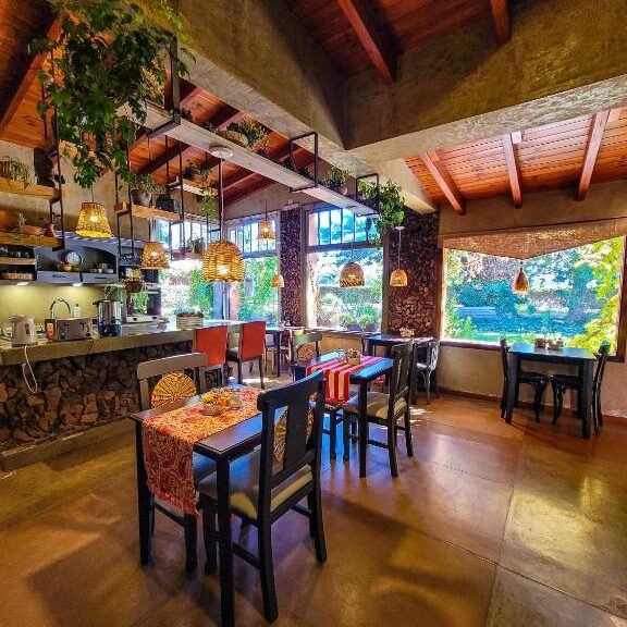 Uma área de jantar aconchegante do Finca La Saucina Casa de Campo com teto de madeira, plantas penduradas, janelas grandes e mesas com toalhas coloridas.