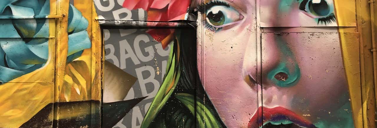 Mural colorido de arte de rua com o rosto surpreso de uma mulher com olhos grandes e flores no cabelo em uma parede texturizada. A imagem ilustra o post sobre Free tour alternativo por Atenas.