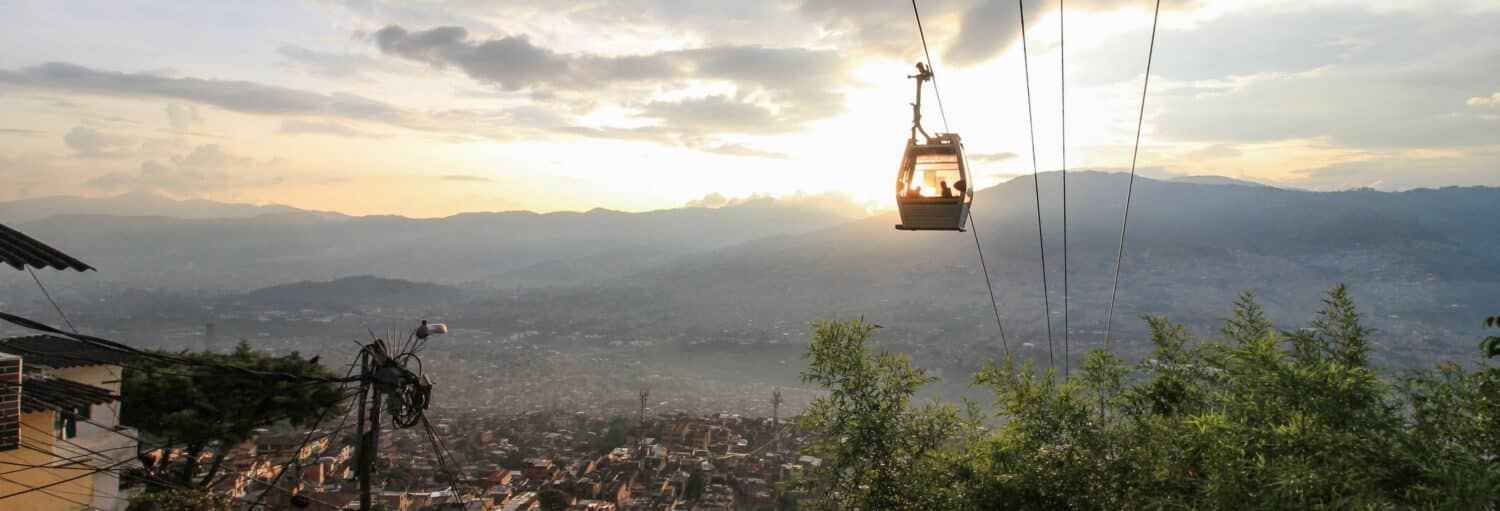 Um teleférico passa por cima de uma paisagem urbana ao pôr do sol, com montanhas e um céu nublado ao fundo. Imagem para ilustrar post sobre o free tour pela Comuna 3 Manrique ao entardecer.
