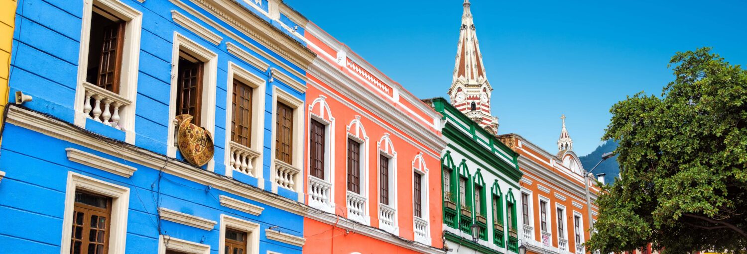 Prédios coloniais coloridos com janelas ornamentadas e uma torre de igreja ao fundo sob um céu azul claro. A imagem ilustra o post sobre Free tour pelo bairro de La Candelaria.