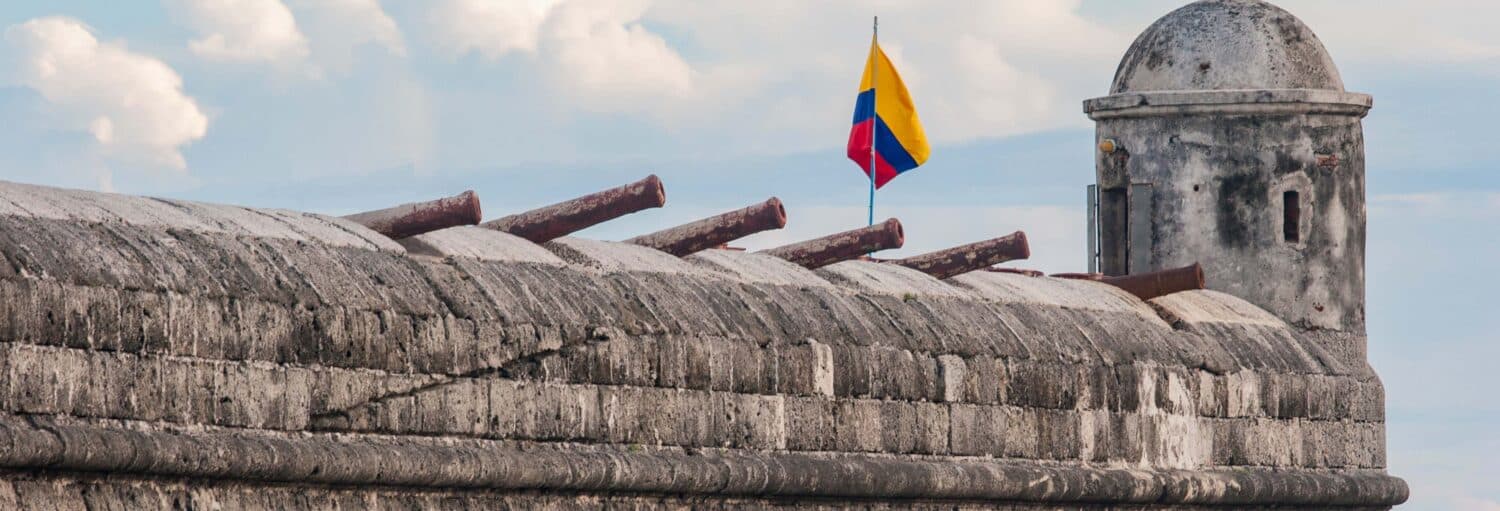 Muro do forte de pedra com canhões, uma torre de vigia e a bandeira colombiana hasteada em um céu nublado. Imagem para ilustrar post sobre o free tour pelo Castelo de San Felipe de Barajas.
