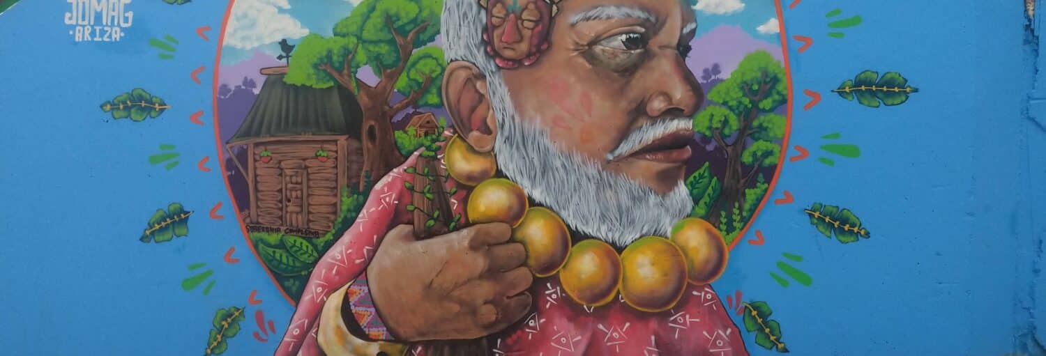 Um mural de um homem idoso com barba branca, usando um grande colar de contas, em um fundo azul com árvores. Imagem para ilustrar post sobre o free tour pela Comuna 13.