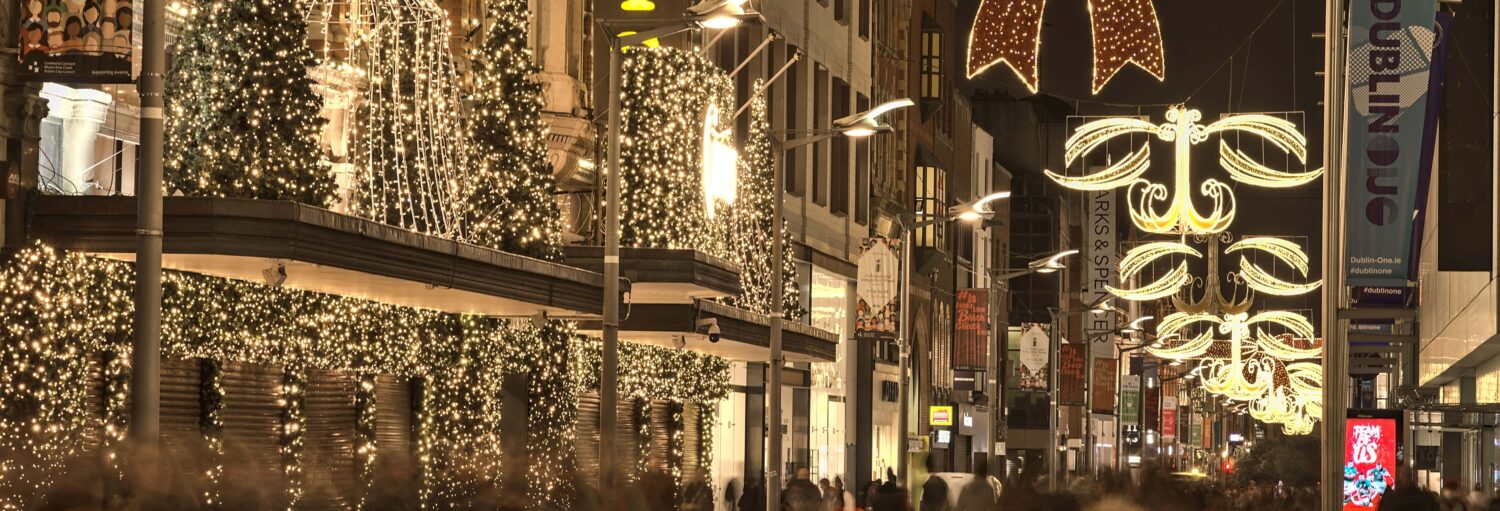 Uma rua da cidade à noite, decorada com luzes de Natal e letreiros luminosos, com prédios e lojas visíveis. A foto é capa do post do Free tour natalino por Dubin.