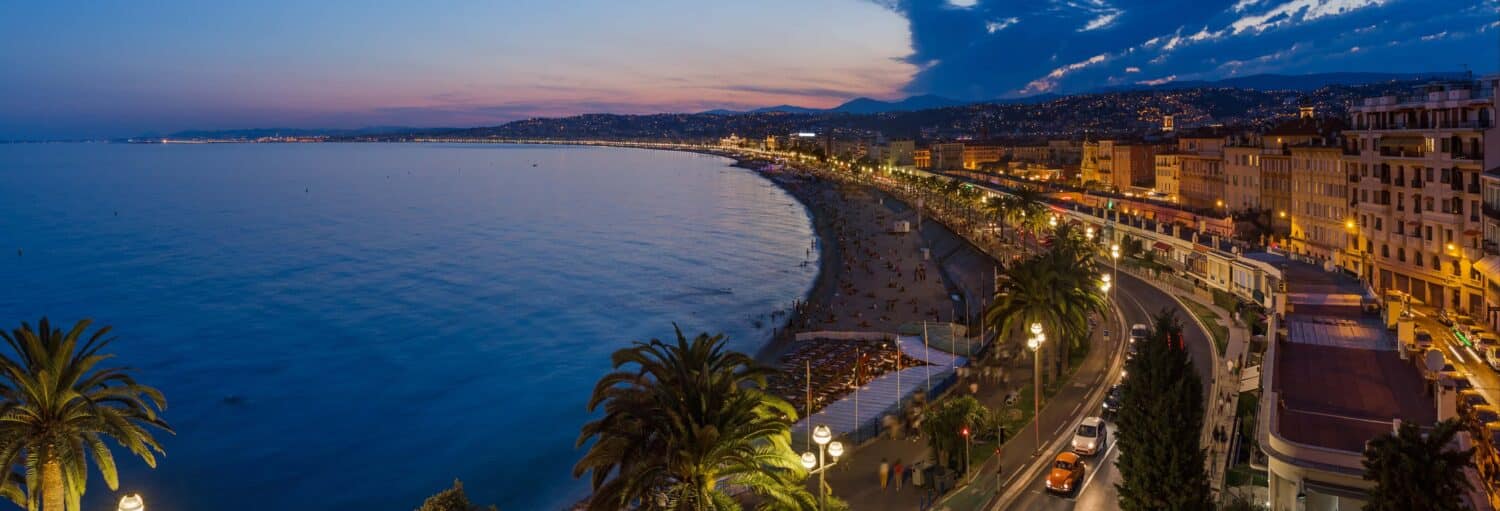 Paisagem urbana litorânea ao anoitecer com uma linha costeira curva, palmeiras, edifícios, postes de luz e colinas distantes sob um céu azul. - Free tour noturno por Nice