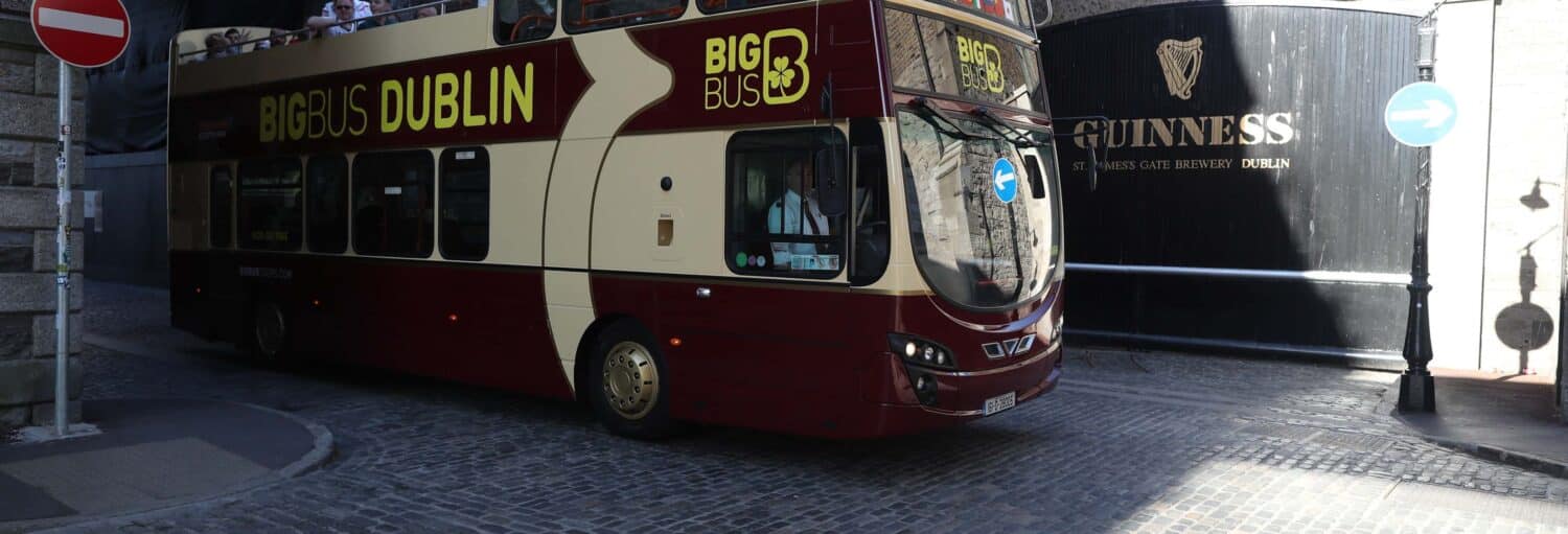 Um ônibus de turismo de dois andares da Big Bus Dublin passa pela entrada da cervejaria Guinness em uma rua de paralelepípedos. A foto é capa do post do Guinness Storehouse + ônibus turístico de Dublin.