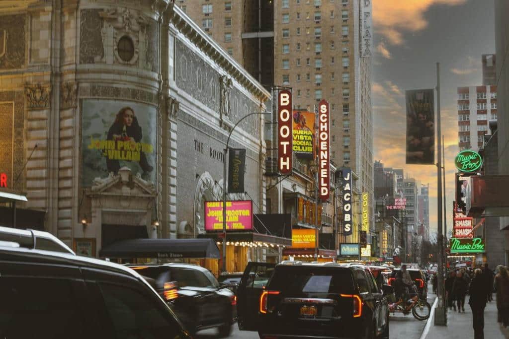 Cena de rua da Broadway ao pôr do sol com carros, pessoas e letreiros iluminados de teatros, incluindo Booth e Schoenfeld.