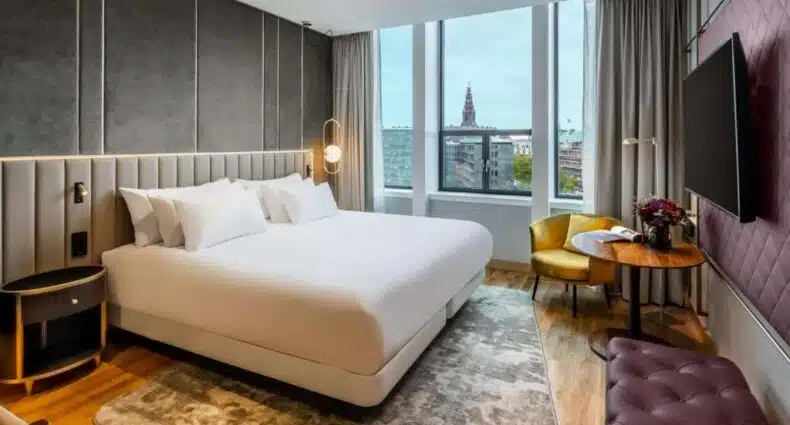 Quarto do hotel NH Collection Copenhagen moderno com uma cama grande, mesa e cadeiras de canto, TV na parede e janelas grandes com vista para a paisagem urbana. Ilustra onde ficar em Copenhague.