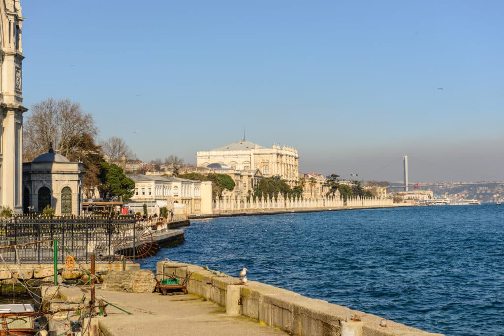 Um calçadão à beira-mar com edifícios históricos, água azul calma e uma ponte suspensa à distância sob um céu claro. Imagem de Beşiktas para ilustrar post sobre onde ficar em Istambul.