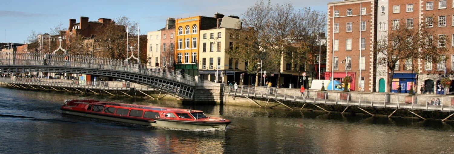 Um barco vermelho viaja em um rio ao lado de edifícios coloridos e uma ponte de pedestres em um dia ensolarado. A foto é capa do post do Passeio de barco por Dublin.