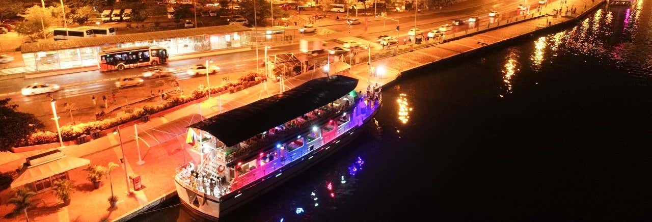 Um barco bem iluminado atracado à beira de um rio à noite, com tráfego e luzes de rua visíveis na estrada adjacente. Imagem para ilustrar post sobre o passeio de barco com espetáculo e jantar por Cartagena.