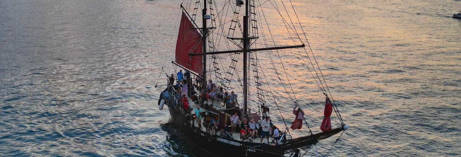 Um grande veleiro com velas vermelhas transporta pessoas por águas calmas ao pôr do sol. Imagem para ilustrar post sobre o passeio de barco pirata ao entardecer.