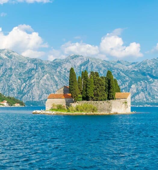 Uma pequena ilha com uma construção de pedra e árvores fica em uma baía azul, cercada por montanhas e nuvens espalhadas, durante o Passeio de barco privado pela baía de Kotor.