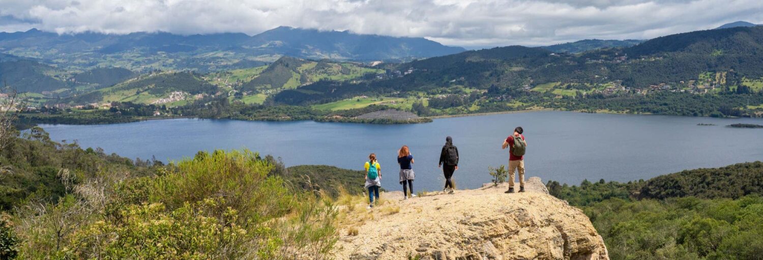 Quatro pessoas estão em um afloramento rochoso com vista para um lago e montanhas verdes sob um céu nublado. A imagem ilustra o post sobre Tour pelas montanhas de La Calera.