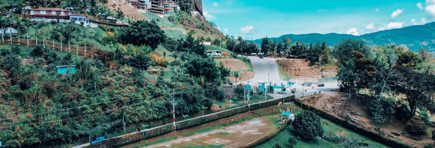 Uma pequena estrada faz curvas em uma área verde e montanhosa com casas e árvores espalhadas sob um céu azul brilhante. Imagem para ilustrar post sobre o passeio de helicóptero pela Comuna 13.