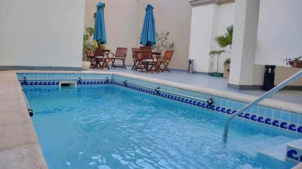 Uma pequena piscina externa do Cóndor Suites Apart Hotel com água azul, cercada por mesas, cadeiras e guarda-sóis azuis em um pátio de azulejos. Imagem para ilustrar post sobre onde ficar em Mendoza.