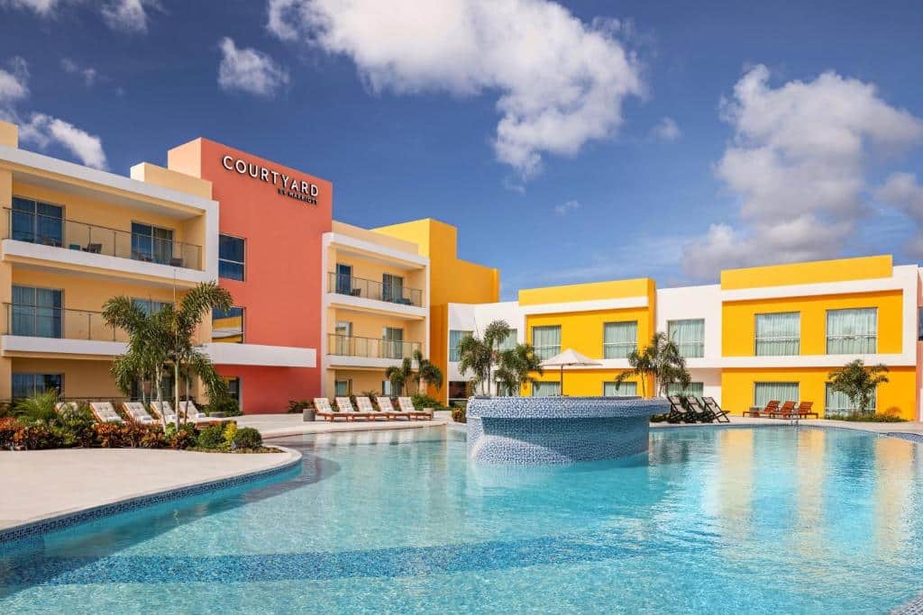 Courtyard by Marriott Curacao com paredes amarelas e laranja, piscina, espreguiçadeiras e palmeiras sob um céu azul.