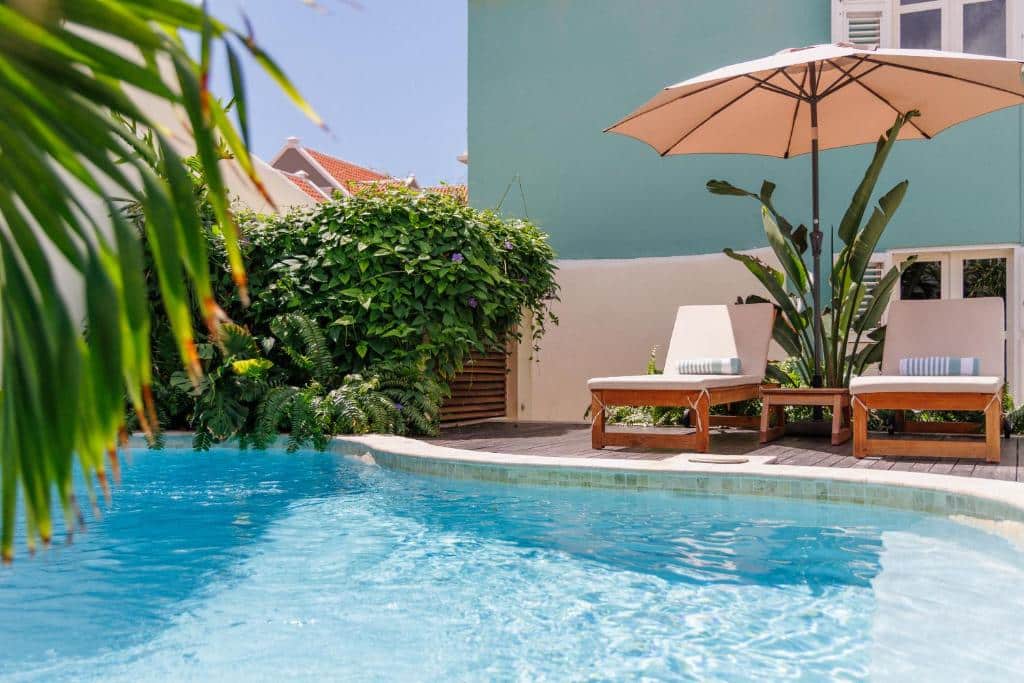 Duas espreguiçadeiras do Pietermaai Oasis e um guarda-chuva estão em um deck de madeira ao lado de uma piscina azul clara perto de um prédio azul claro e plantas verdes.