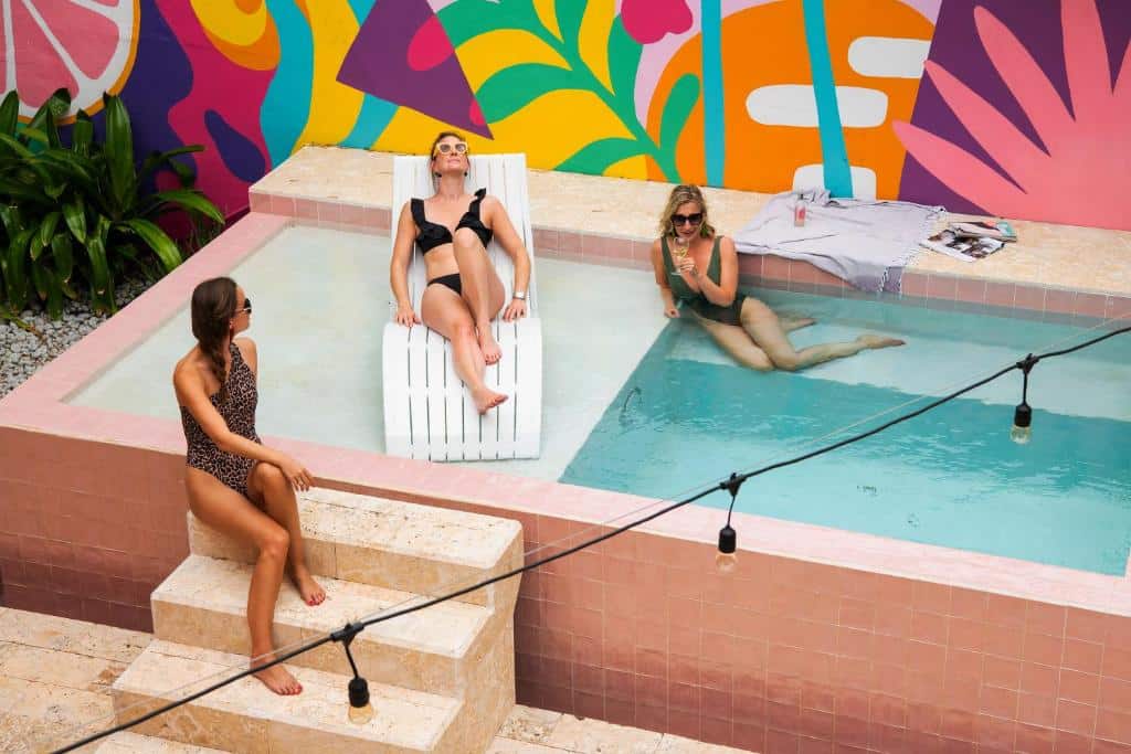 Três mulheres em trajes de banho relaxam em uma pequena piscina do Wynwood Boutique Hotel com um mural colorido ao fundo e luzes de corda no alto.
