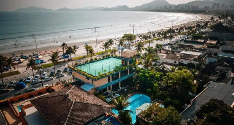 Vista aérea da Praia da Enseada com edifícios e palmeiras ao longo da orla, quadra de tênis no primeiro plano e pessoas aproveitando o dia ensolarado na areia. Um dos edifícios é a Pousada Stand Up! Guarujá