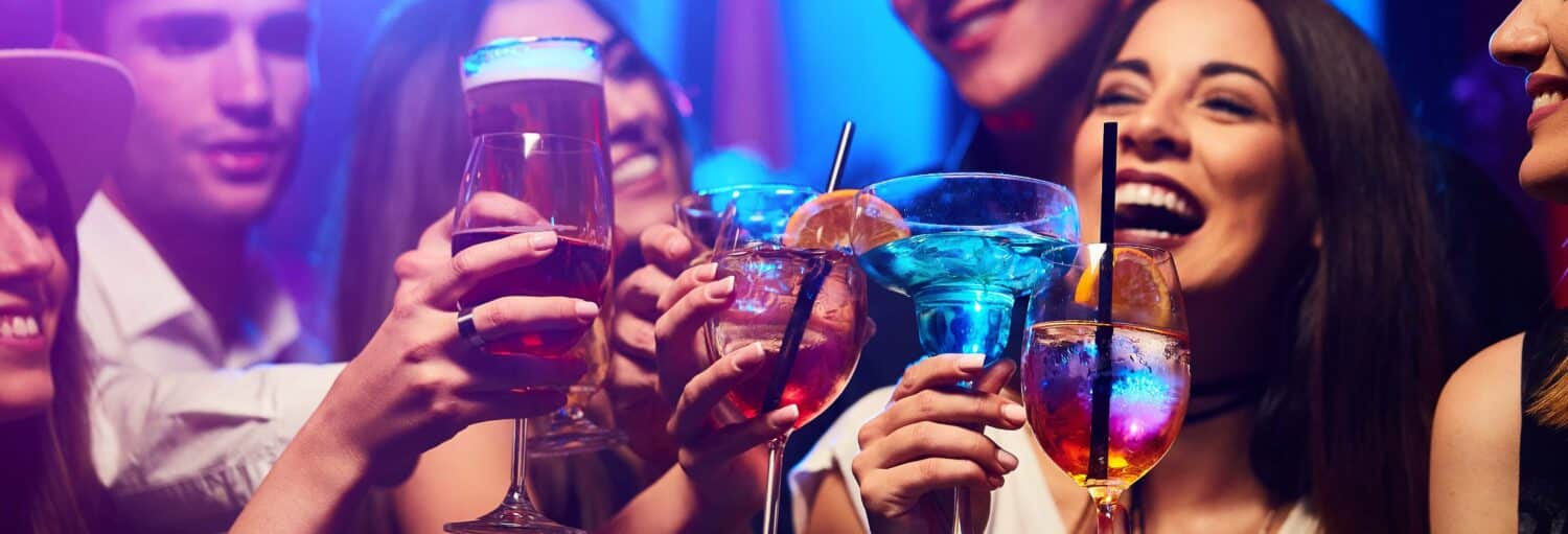 Um grupo de pessoas levantando coquetéis e bebidas coloridas enquanto sorriem e comemoram em um ambiente animado de bar ou clube. - Pub Crawl. Tour de festa por Marselha!