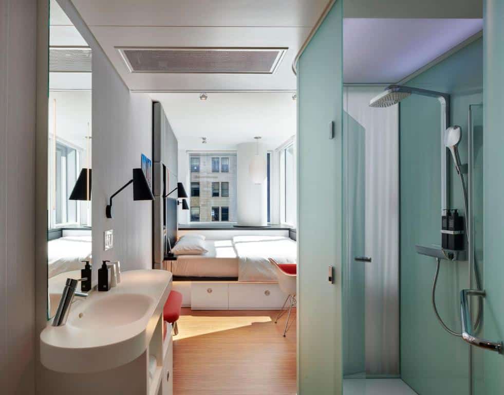 Um quarto moderno no CitizenM Chicago Downtown com uma cama, pia branca, luminárias de parede, chuveiro de vidro e janelas grandes voltadas para um prédio da cidade, para representar hotéis em Chicago.