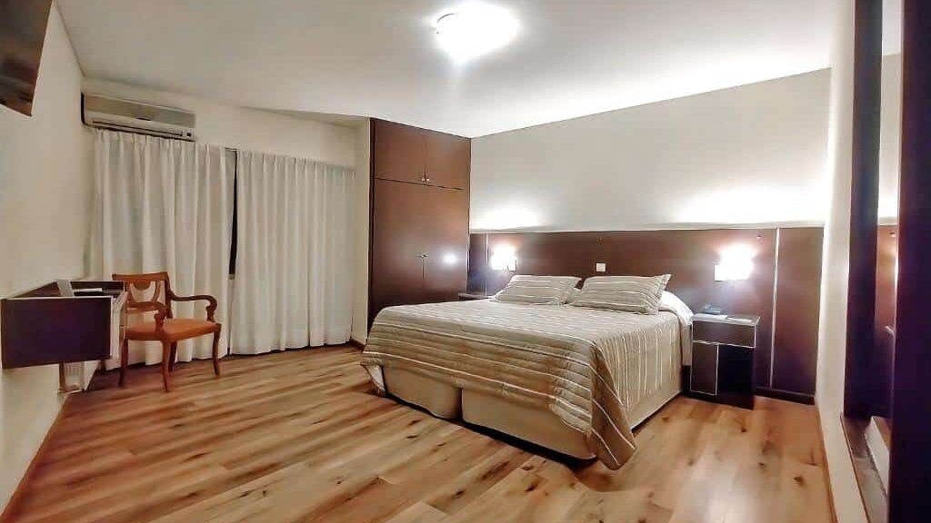 Um quarto espaçoso do Cóndor Suites Apart Hotel com uma cama grande, piso de madeira, duas cadeiras, um guarda-roupa e iluminação suave.