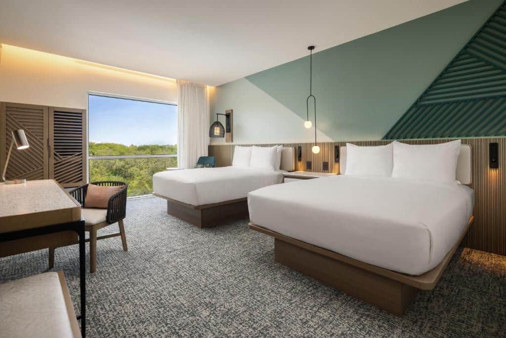 Quarto do Courtyard by Marriott Curacao com duas camas de casal, uma escrivaninha com cadeira, janela grande, decoração moderna e parede com detalhes em verde.