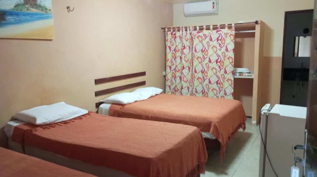 Um quarto com três camas de solteiro cobertas com cobertores laranja, um armário com cortinas, ar-condicionado e um pequeno frigobar.