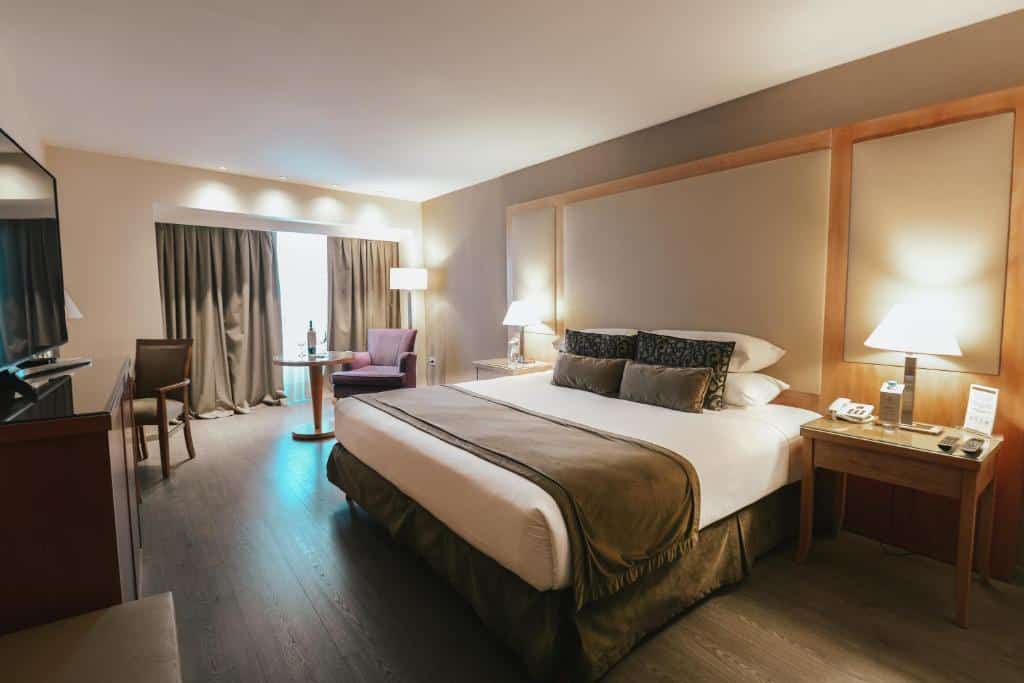 Um quarto do DiplomaticHotel com uma cama grande, duas mesas de cabeceira, luminárias, cadeiras, uma mesa, cortinas e uma TV em um suporte.