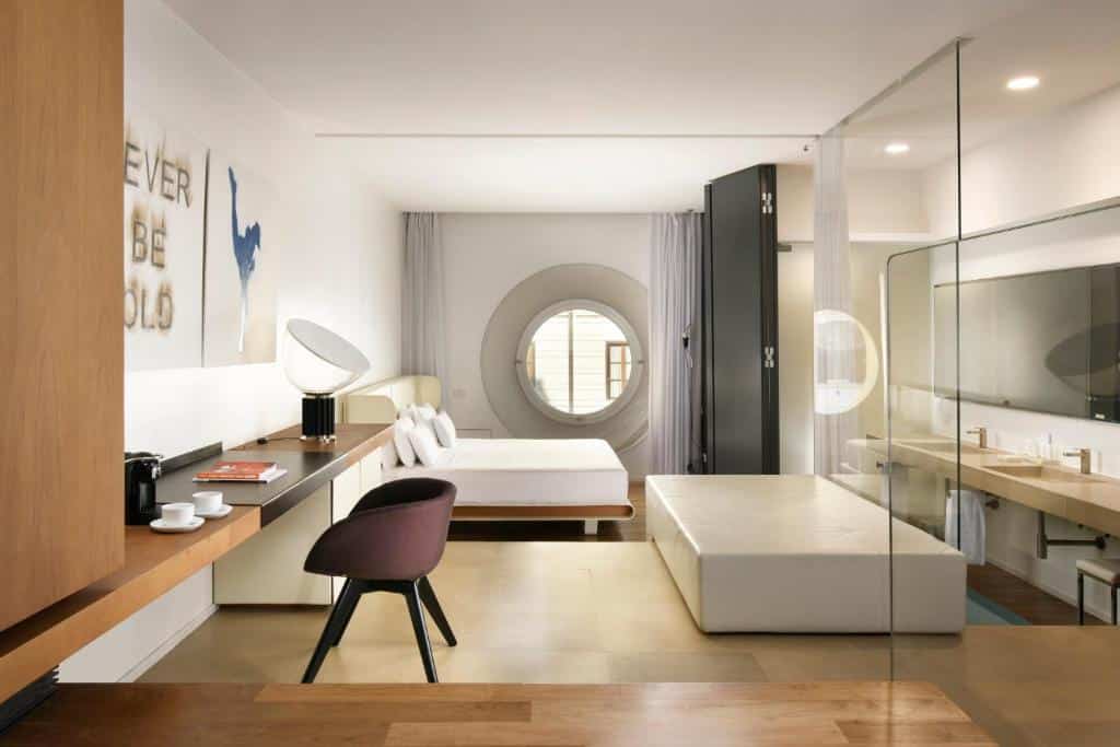 Quarto do Palazzo Montemartini Rome, A Radisson Collection Hotel com janela redonda, cama de casal, escrivaninha, cadeira e área de banheiro com paredes de vidro.