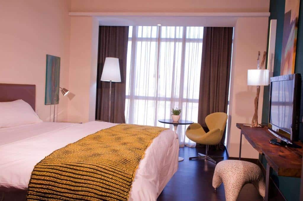 Um quarto do Esplendor by Wyndham Mendoza com uma cama, cobertor amarelo, cadeira, TV, mesa pequena, luminária e uma janela grande com cortinas.