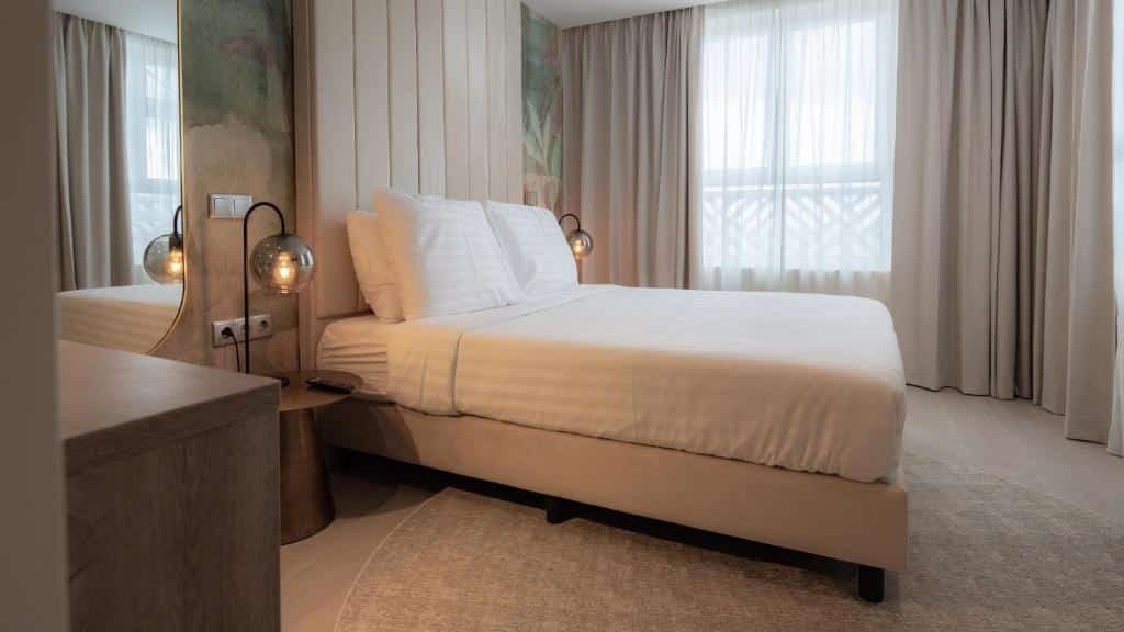 Uma cama bem arrumada do Harbor Hotel & Casino Curacao com lençóis brancos em um quarto de hotel moderno com decoração neutra e janelas grandes com cortinas.